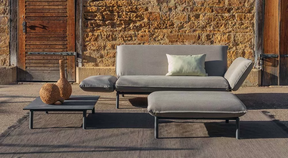 Fischer Möbel Luna Loungegruppe in Grau auf einer Terrasse mit rustikaler Steinwand im Hintergrund, bestehend aus Sofa, Hocker und Tisch.