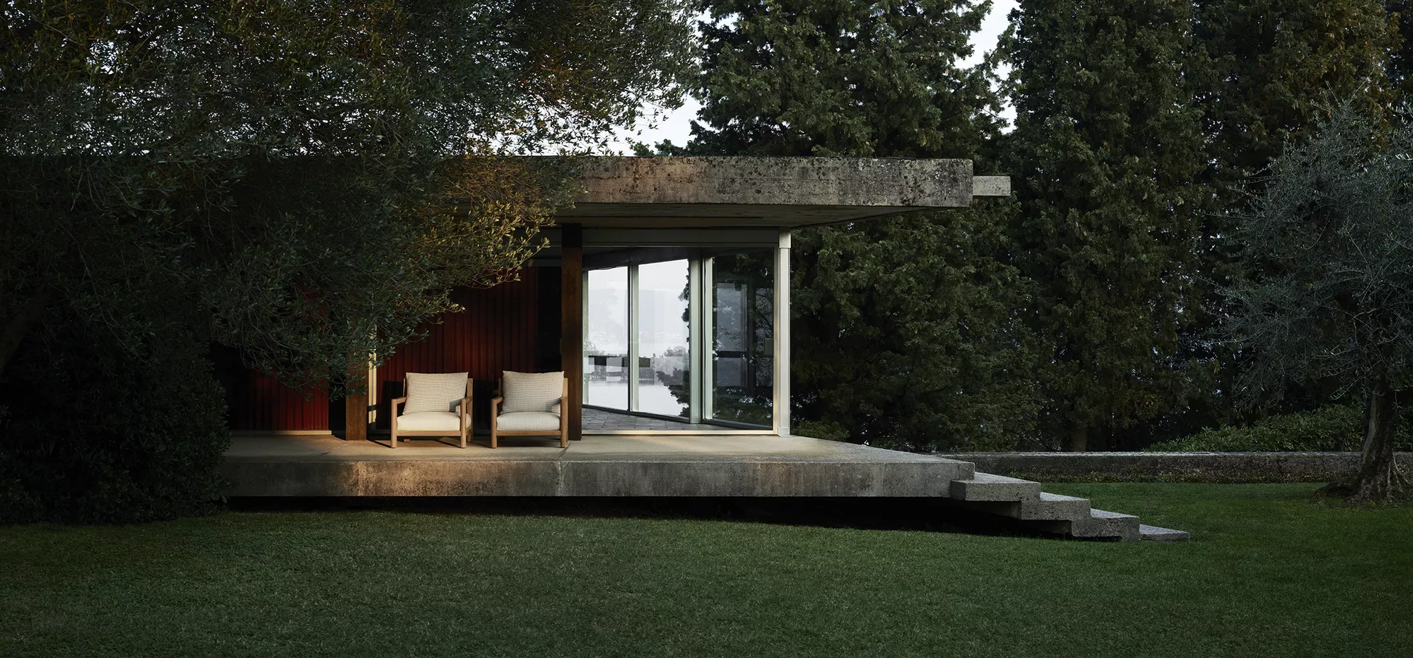 Zwei B&B Italia Pablo Outdoor Lounge Sessel stehen auf einer Terrasse vor einem modernen Haus umgeben von Bäumen in einer ruhigen Umgebung.