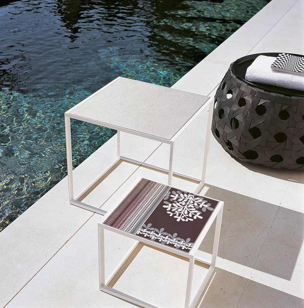 Zwei B&B Italia Canasta Outdoor Beistelltische stehen nebeneinander am Pool einer hat ein dekoratives Muster der andere ist schlicht.