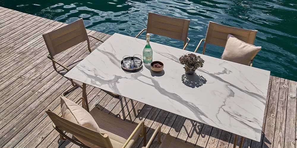 Weishäupl Sosta Tisch in Marmoroptik mit vier Holzstühlen auf einem Steg am Wasser, stimmungsvolles Outdoor-Setting.