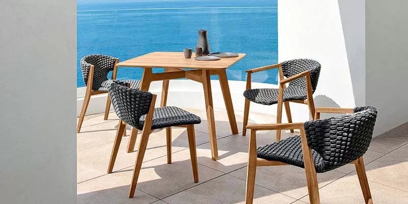 Ethimo Knit Outdoor Essgruppe in Naturholz und Dunkelgrau auf einer Terrasse mit Meerblick bei sonnigem Wetter.