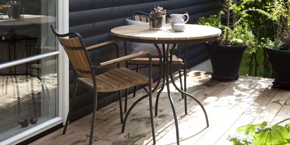 Gartenszene mit einem runden Holztisch und Stuhl auf einer Holzterrasse, umgeben von Pflanzen und einem schwarzen Haus im Hintergrund.