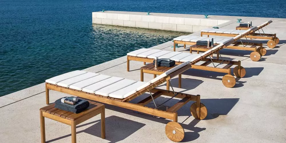 Vier Varaschin Barcode Gartenmöbel in Holzoptik mit weißen Polstern stehen auf einer Terrasse am Wasser.