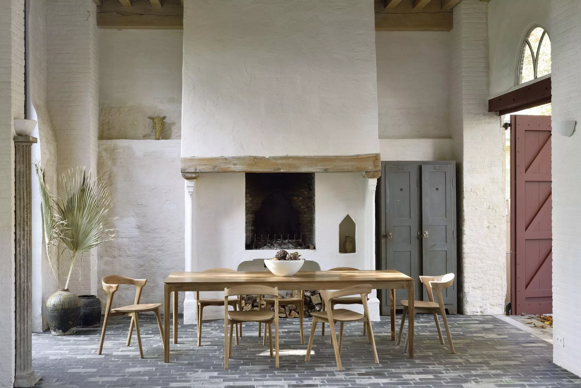 Ethnicraft Bok Tisch aus Teakholz in einem rustikalen Raum mit Steinboden und Kamin, umgeben von passenden Stühlen.