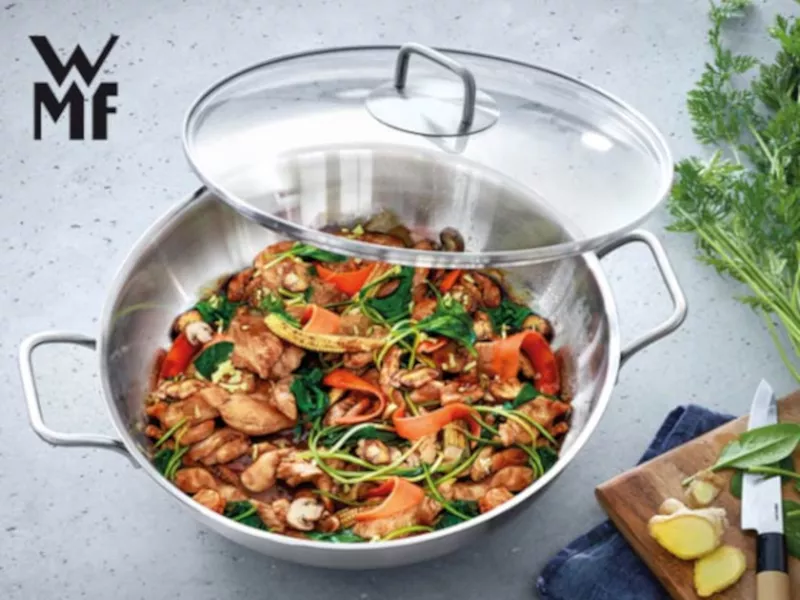 WMF Wok-Pfanne, 28 cm, aus Edelstahl, mit Glasdeckel, gefüllt mit buntem Gemüse und Hähnchen, auf einem Holztisch.