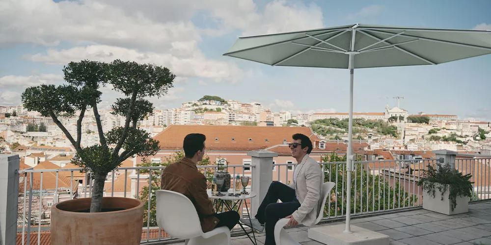 Zwei Personen sitzen unter einem Glatz Twist Sonnenschirm in Hellgrün auf einer Terrasse mit Stadtblick im Hintergrund.