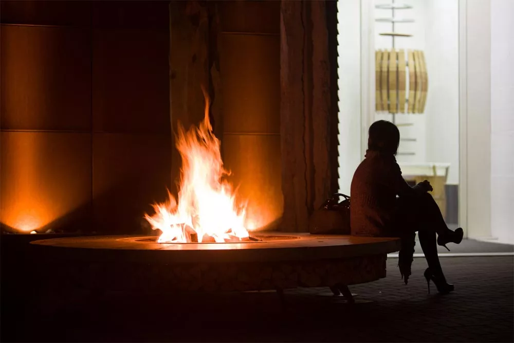 Eine Person sitzt neben der AK47 Ercole Beton Stahl Feuerstelle im Freien während die Flammen in die Höhe schlagen und eine warme Atmosphäre schaffen.