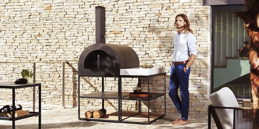 Ein Mann lehnt an einem Roshults BBQ Wood Oven in Schwarz vor einer Steinmauer, umgeben von modernen Gartenmöbeln.