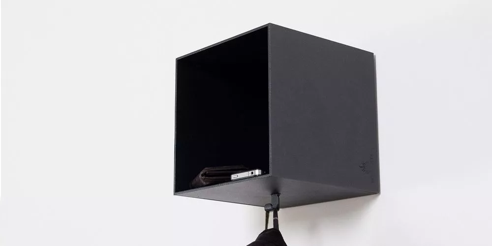 Schwarzes Roshults Regal aus Metall, an einer weißen Wand montiert, mit einem Haken darunter und einem Smartphone im Inneren.