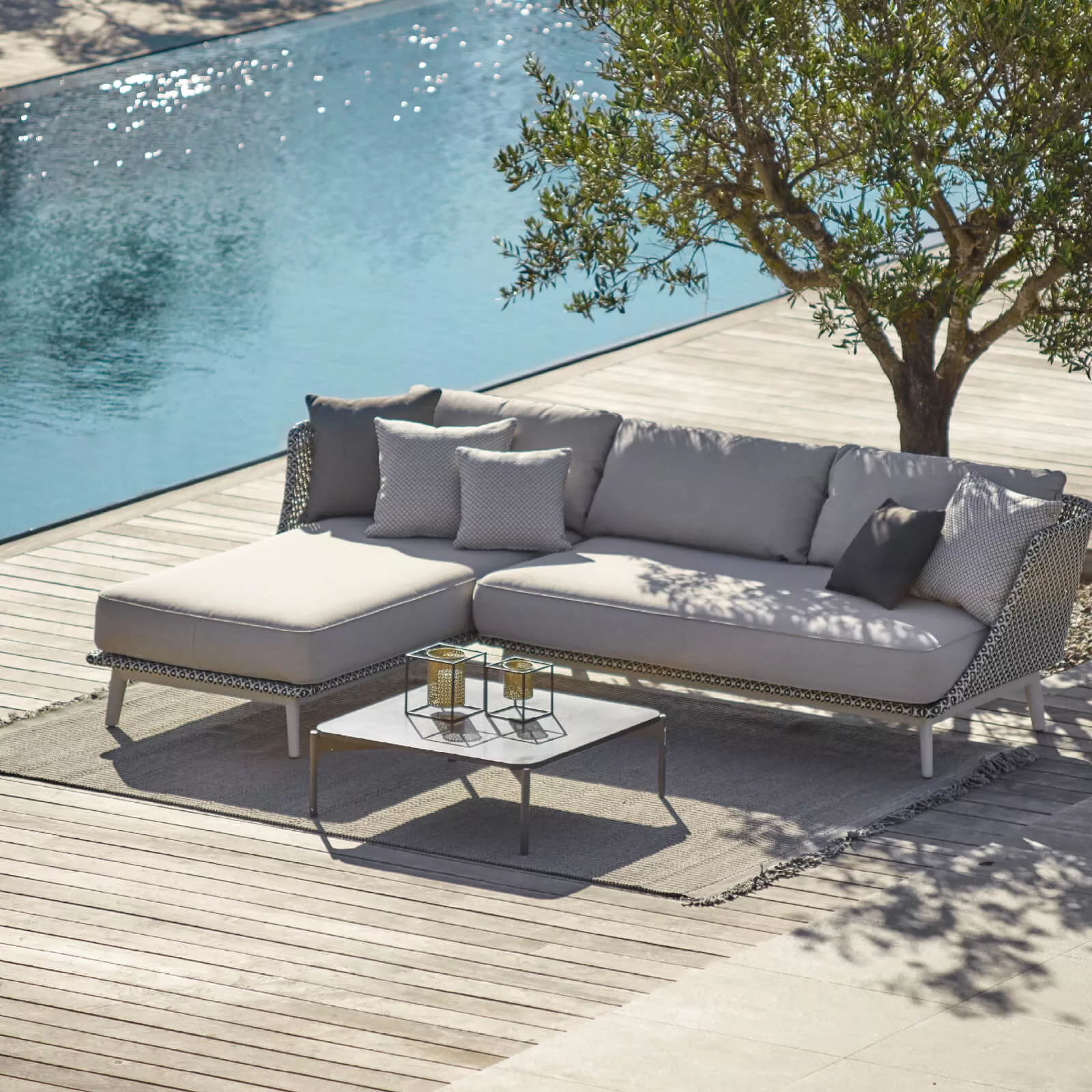 Dedon Mbarq Lounge Sofa in Grau mit Kissen und Couchtisch auf einer Terrasse neben einem Pool und einem Olivenbaum in einer entspannten Atmosphäre.