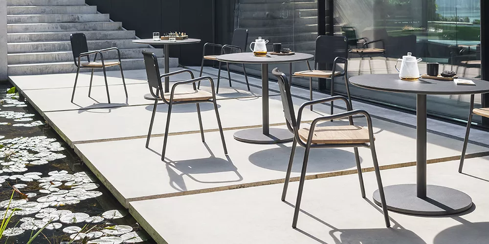 Außenbereich mit Roda Kollektion Butten in Schwarz, moderne Stühle und Tische auf heller Terrasse neben einem Teich, sonnige Atmosphäre.