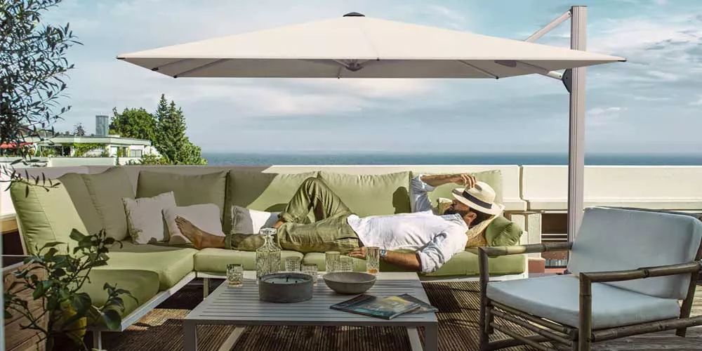 Ein Mann entspannt auf einem Sofa unter einem weißen Glatz Ambiente Nova Sonnenschirm auf einer Terrasse mit Meerblick.