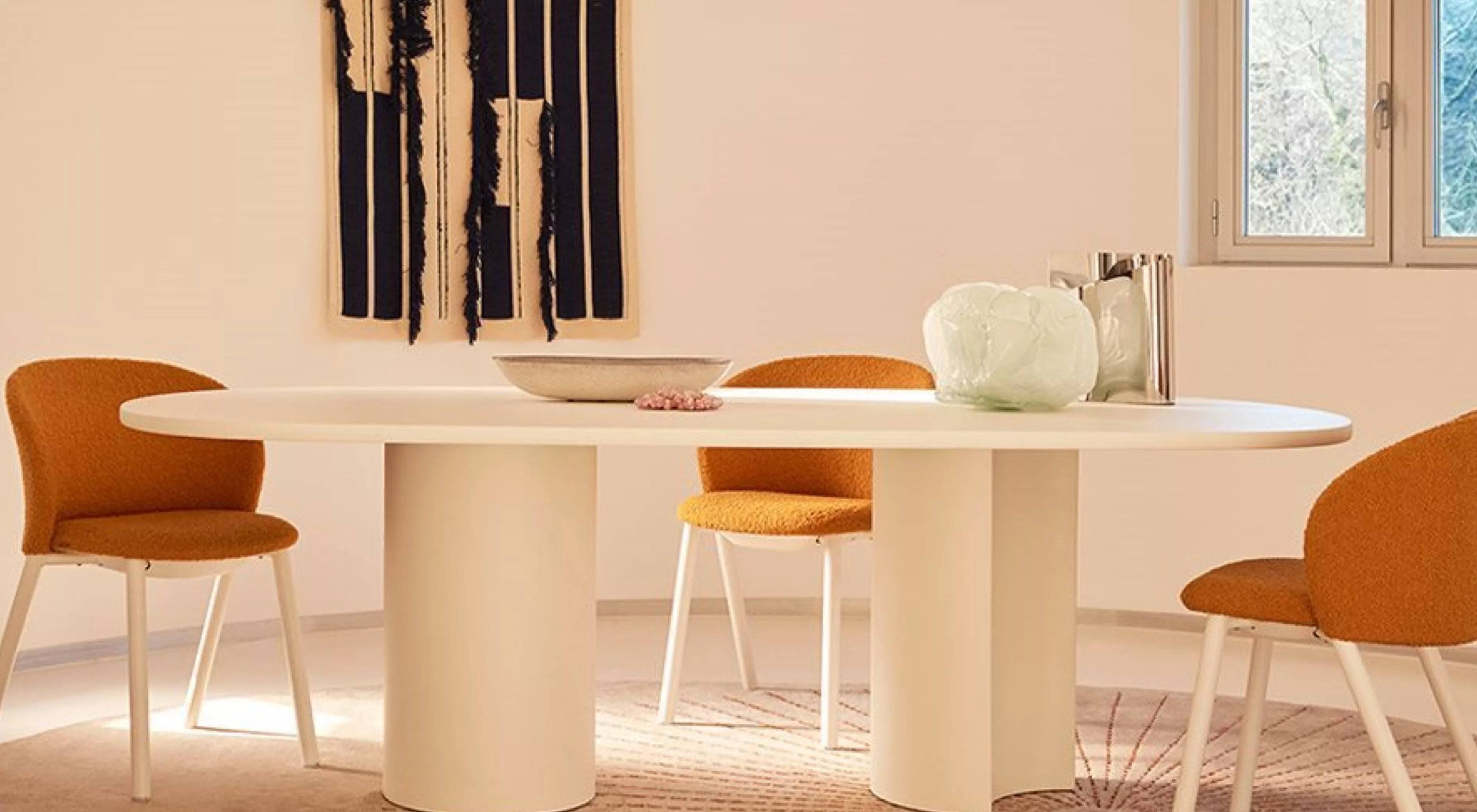 Ovaler weißer Tisch mit orangefarbenen Stühlen in modernem Wohnzimmer, Wandkunst im Hintergrund, Tageslicht durch Fenster.
