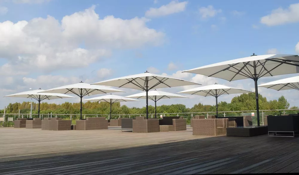 Bahama Jumbrella Sonnenschirme in Weiß auf einer Holzterrasse mit Sitzgruppen, vor einem Hintergrund aus Bäumen und blauem Himmel.