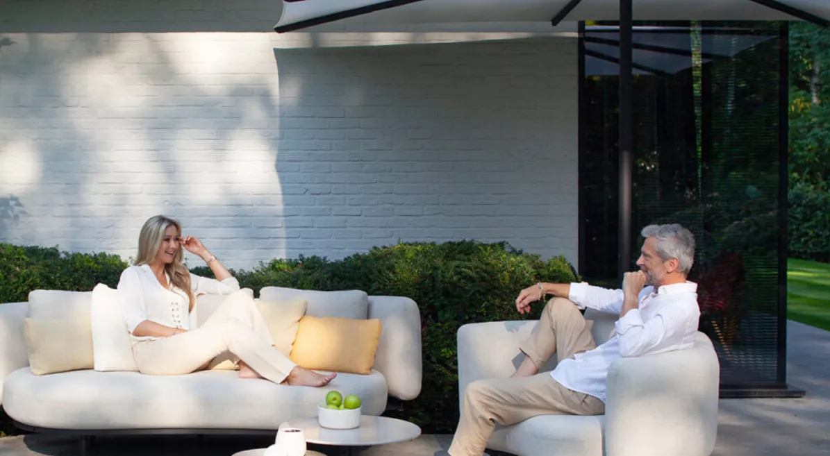 Zwei Personen entspannen auf weißen Dedon Sofas im Garten, umgeben von grünen Büschen und einem modernen Gebäude im Hintergrund.