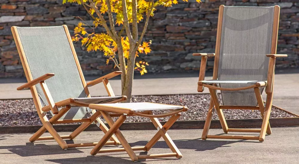 Zwei fischer möbel Tennis Lounge-Sessel und ein Hocker aus Holz und grauem Stoff auf einer Terrasse vor einer Steinmauer und einem Baum.