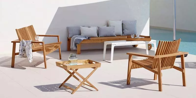 Cane-line Amaze Loungegruppe aus Teakholz auf heller Terrasse mit Pool im Hintergrund, bestehend aus zwei Sesseln, Tisch und Sofa.