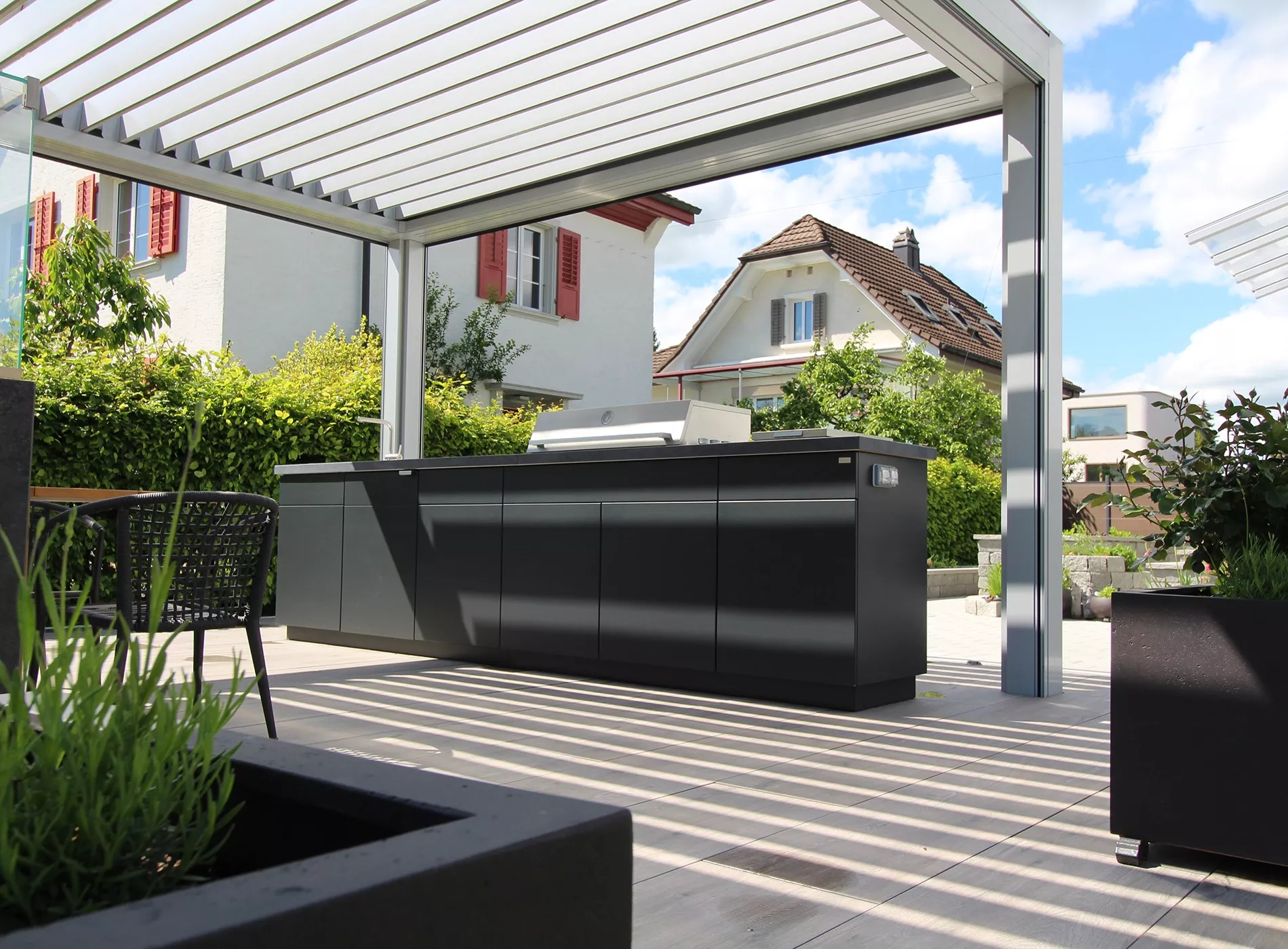 Cubic Outdoor Kitchen in Anthrazit auf einer Terrasse mit modernem Lamellendach und Blick auf ein Haus im Hintergrund.