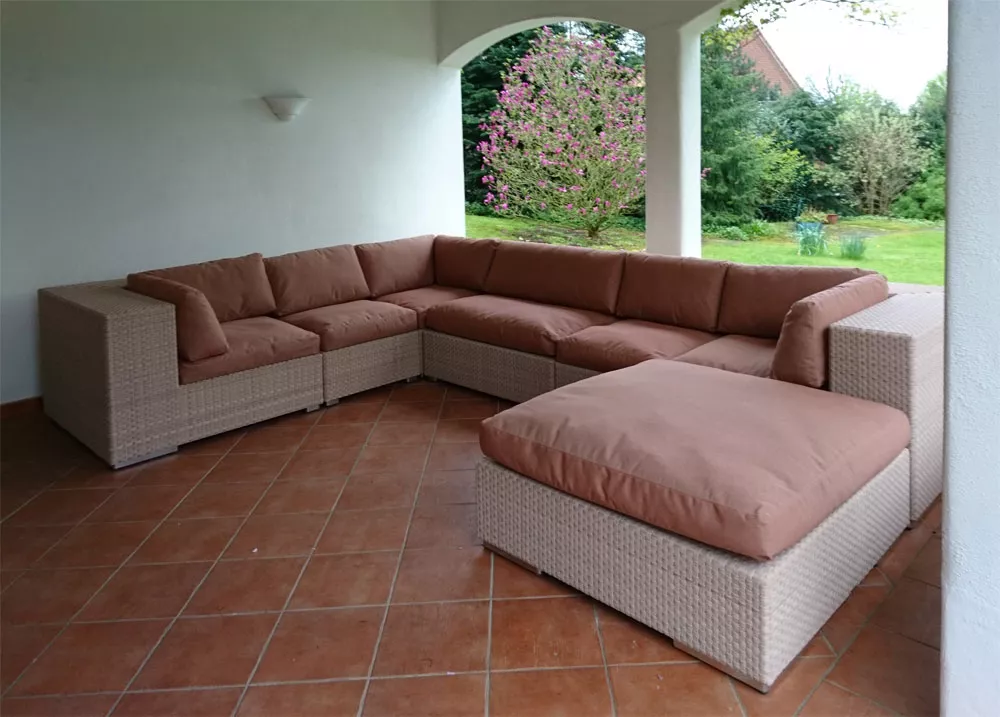 Dedon Lounge mit beigen Polstern steht auf einer überdachten Terrasse mit Blick auf einen grünen Garten und blühenden Baum im Hintergrund