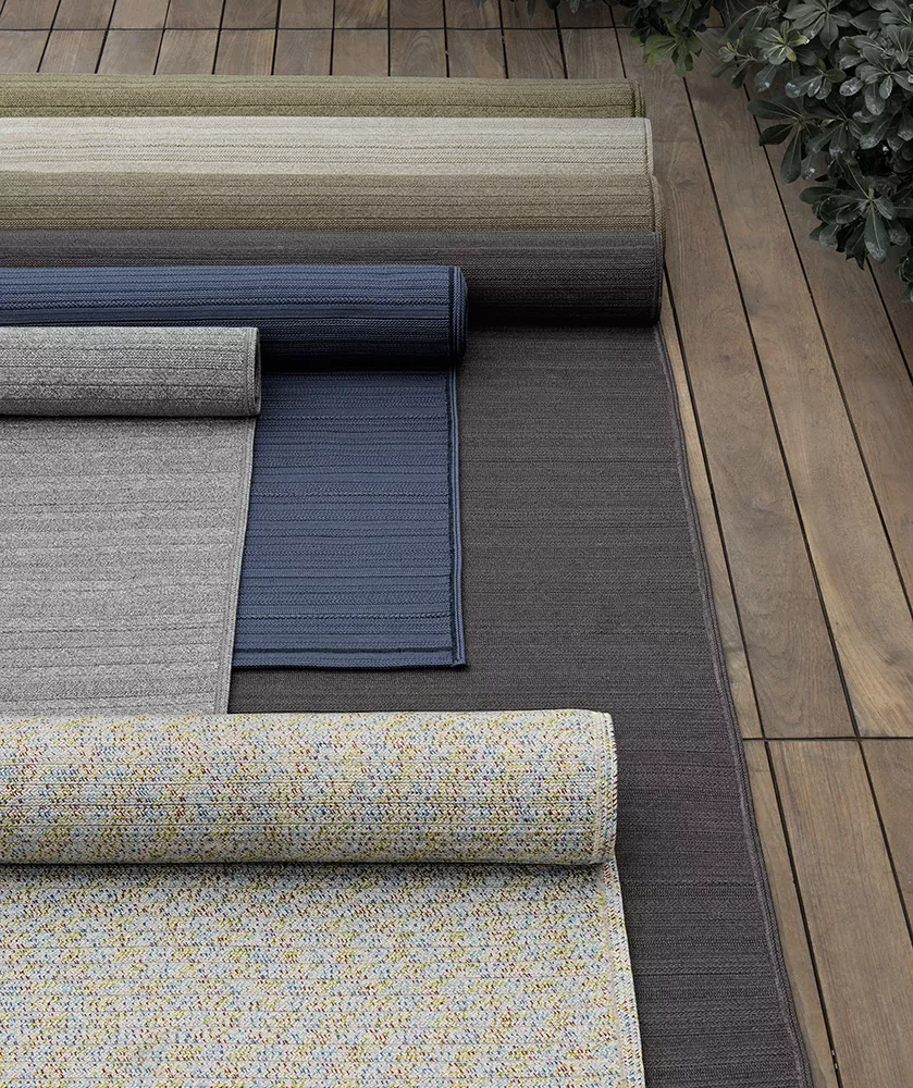 Mehrere B&B Italia Teppiche in verschiedenen Farben und Mustern auf einem Holzboden ausgerollt mit Pflanzen im Hintergrund.