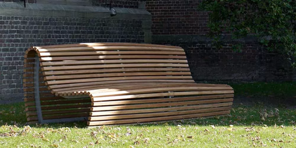 B&B Italia Titikaka Bank aus Holz steht auf einer Wiese vor einer Backsteinmauer im Schatten eines Baumes