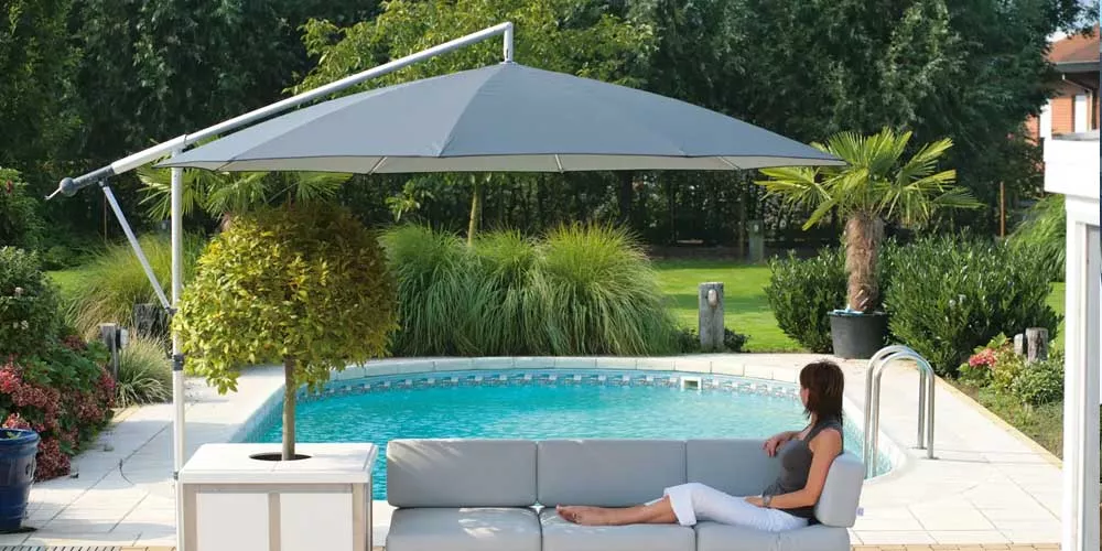 Frau entspannt auf grauem Sofa unter einem grauen May Sonnenschirm Mezzo am Pool, umgeben von grüner Gartenlandschaft.