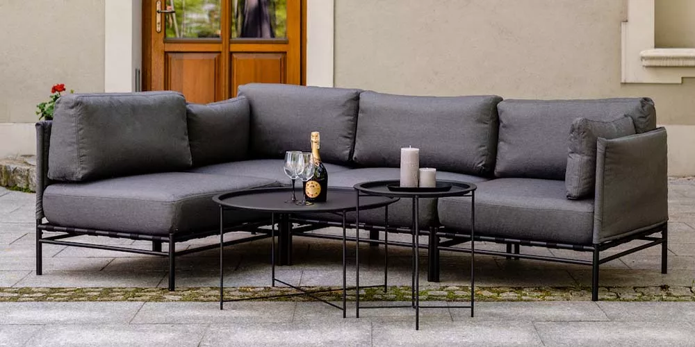 Grau, stilvolles Outdoor-Sofa aus der RM Bern Kollektion mit schwarzem Metallrahmen, abgebildet auf einer Terrasse vor einer Holztür.