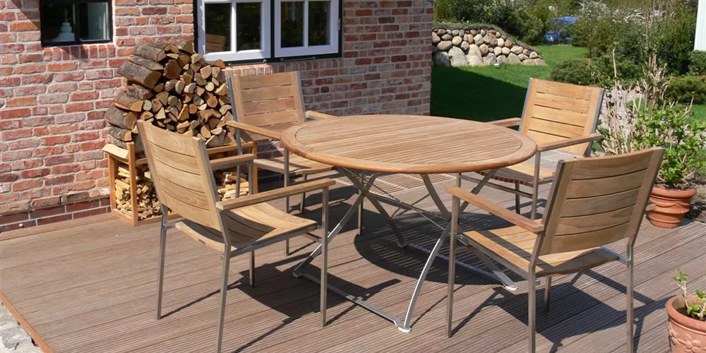 Gartenmöbel-Set von Royal Mirage mit rundem Holztisch und vier Stühlen auf einer Holzterrasse vor einem Backsteinhaus.