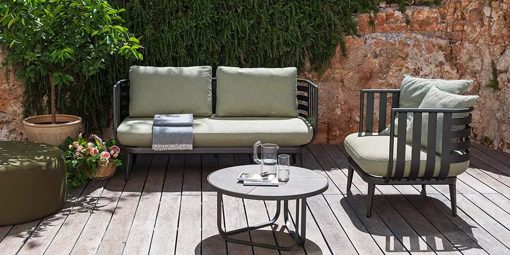 Roda Thea Loungegruppe in Grün auf Holzterrasse vor Steinmauer und Pflanzen, bestehend aus Sofa, Sessel und rundem Tisch, stimmungsvolles Ambiente.