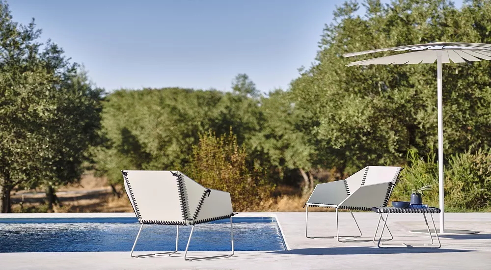 Zwei Textile Club Sessel in Achatgrau stehen auf einer Terrasse neben einem Pool, umgeben von Olivenbäumen und einem Sonnenschirm.