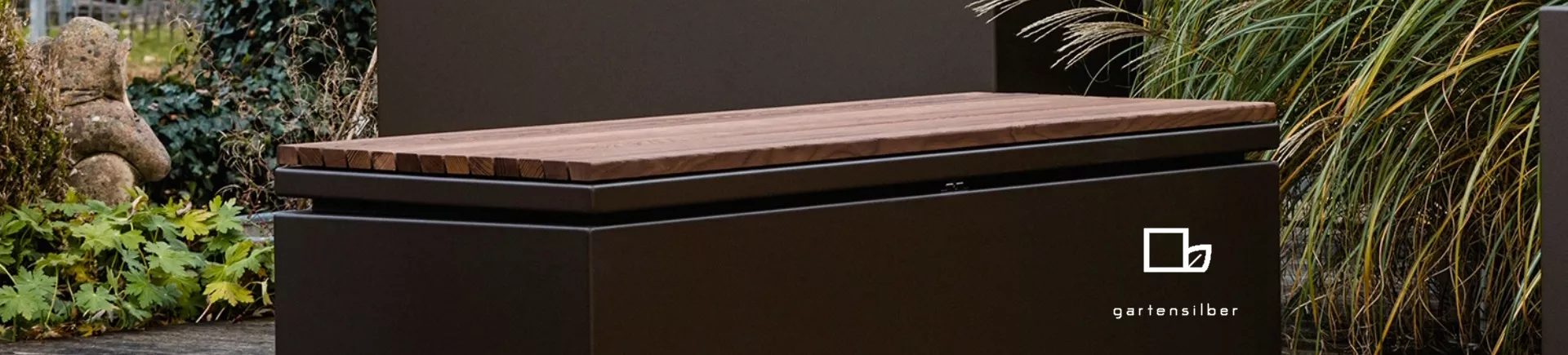 Braune Gartensilber-Bank von Villa Schmidt auf einer Terrasse, umgeben von Pflanzen, mit modernem Design und Holzsitzfläche.