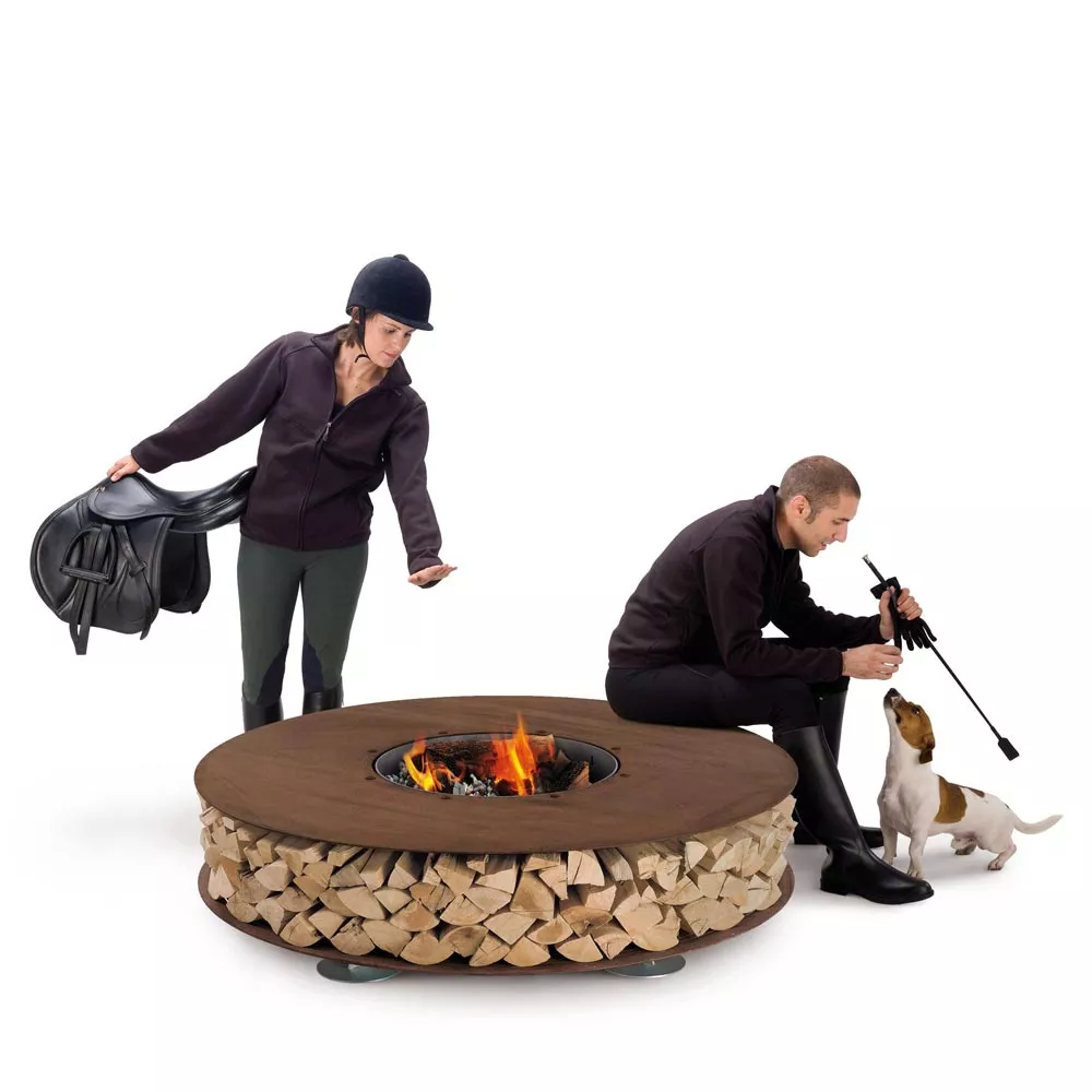 Zwei Personen und ein Hund sitzen und stehen um die AK47 Zero Feuerstelle 145 cm Gartenkamin herum und genießen die Wärme des Feuers.