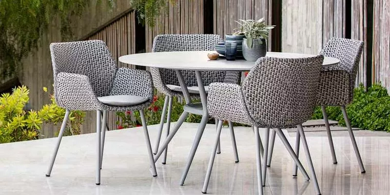 Cane-line Joy Gartenmöbel-Set in Grau auf einer Terrasse mit Pflanzen im Hintergrund, bestehend aus einem runden Tisch und vier Stühlen.