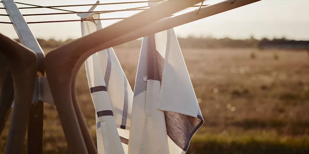 Skagerak Outdoor-Wäschespinne in Braun mit aufgehängten weißen Handtüchern, im Hintergrund eine sonnige Wiesenlandschaft.