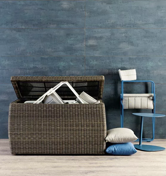 Ethimo Barth Kissentruhe in Braun, 116 cm, vor einer blauen Wand mit Kissen und einem blauen Stuhl im Hintergrund.