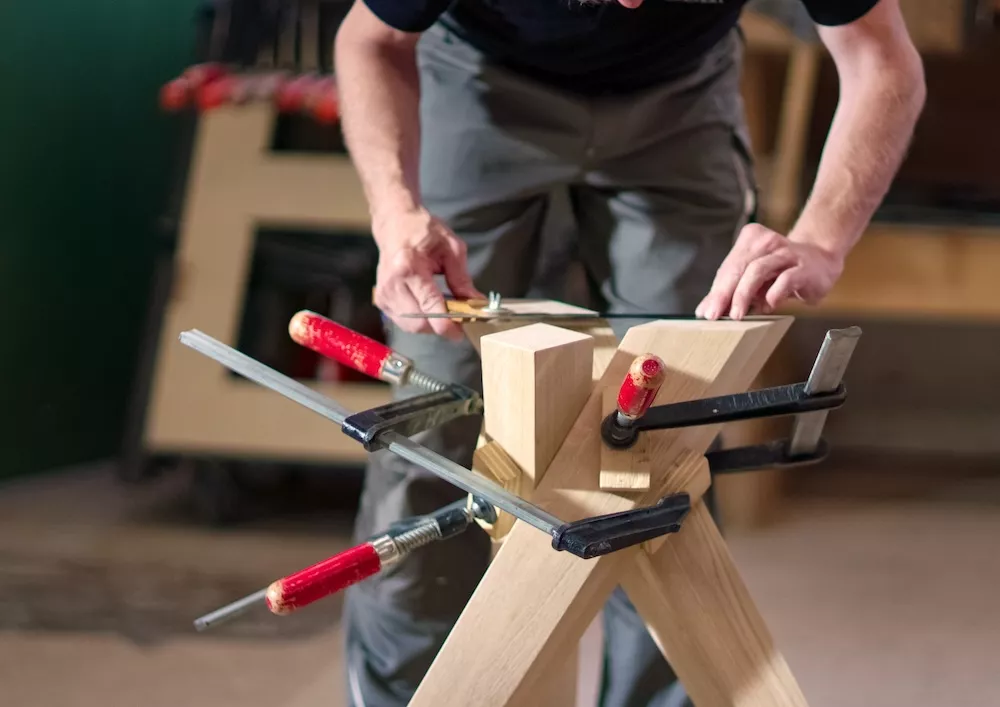 Ein Handwerker bearbeitet ein Holzstück mit einer Feile das mit mehreren Zwingen fixiert ist in einer Werkstatt.