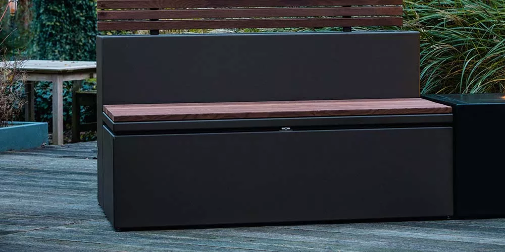 Kissentruhe von Villa Schmidt in Anthrazit mit Holzsitzfläche, auf einer Terrasse im Freien, umgeben von Pflanzen.