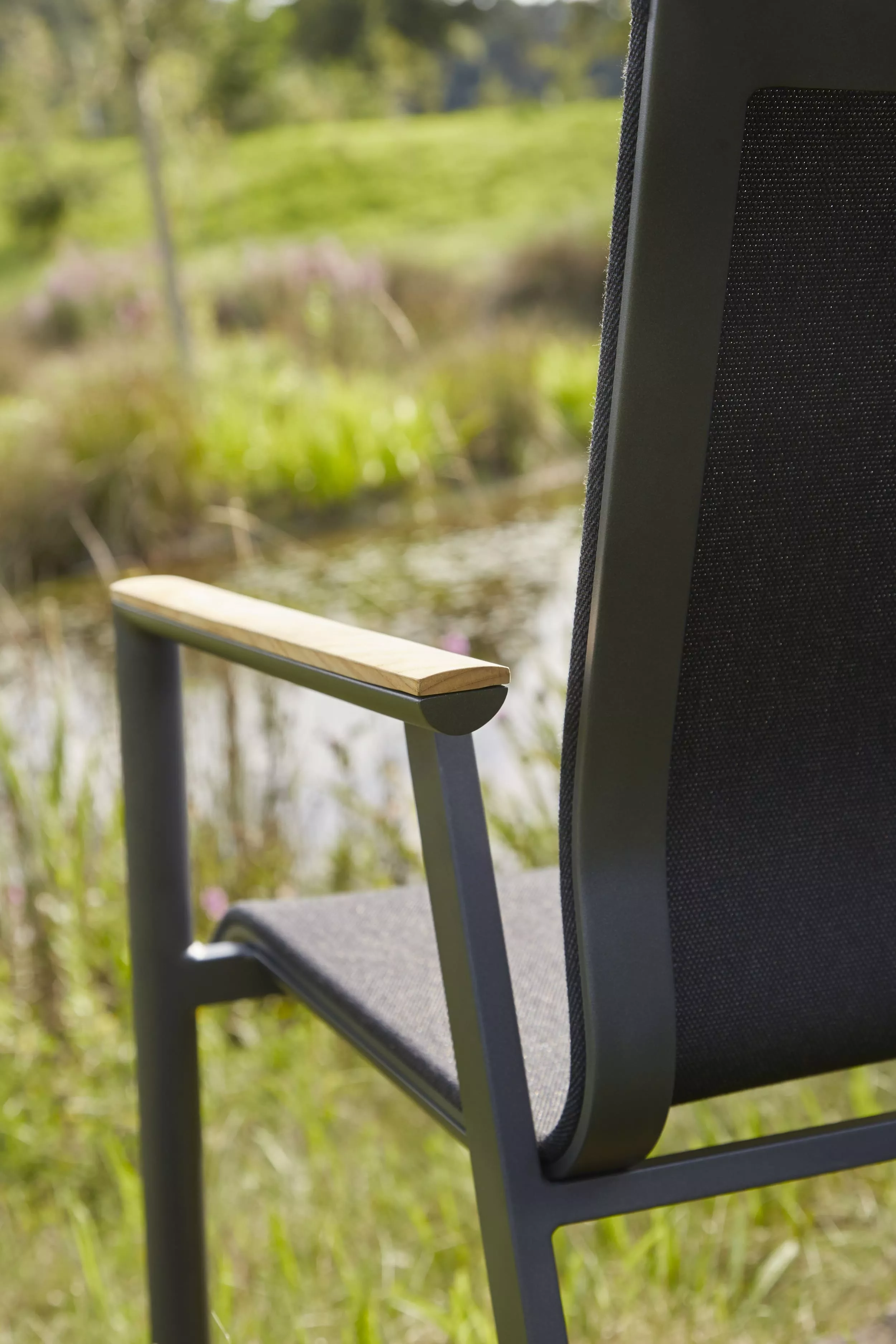 Solpuri Soul Stuhl mit schwarzem Netzgewebe und Armlehnen aus Holz steht in einem grünen Garten vor einem Teich Moodbild