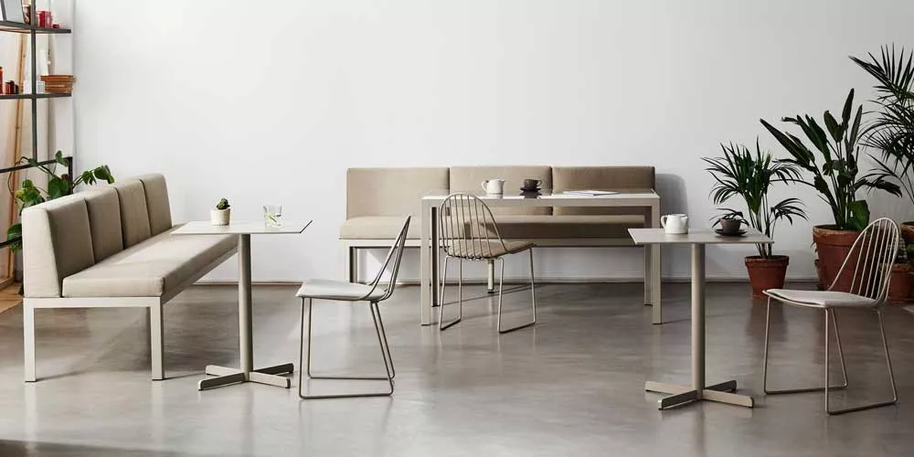 Essgruppe Bivaq NAK65 in Beige, bestehend aus Tisch, Bank und Stühlen, auf Betonboden mit Pflanzen im Hintergrund.