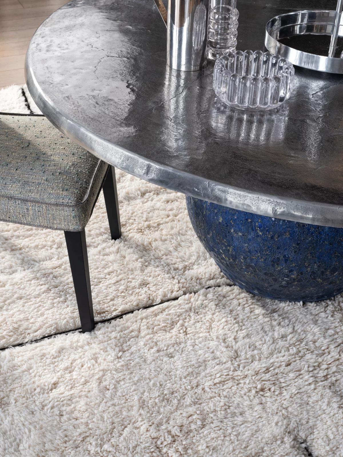 Gervasoni Tisch mit Metallplatte und blauer Basis auf einem flauschigen Teppich mit einem Stuhl und Glasdekorationen Moodbild Wohnzimmer