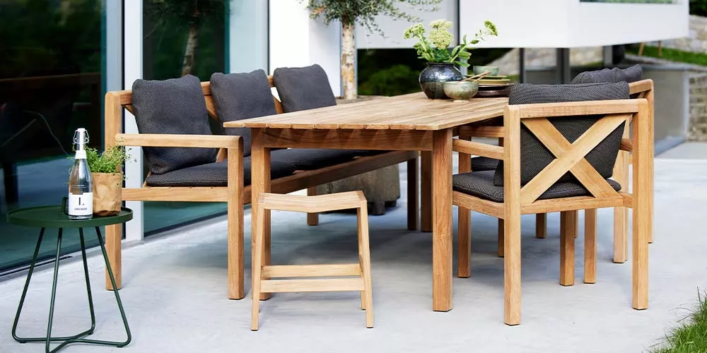 Cane-line Grace Kollektion, Holztisch und Stühle mit grauen Kissen, auf einer Terrasse vor einem modernen Gebäude.