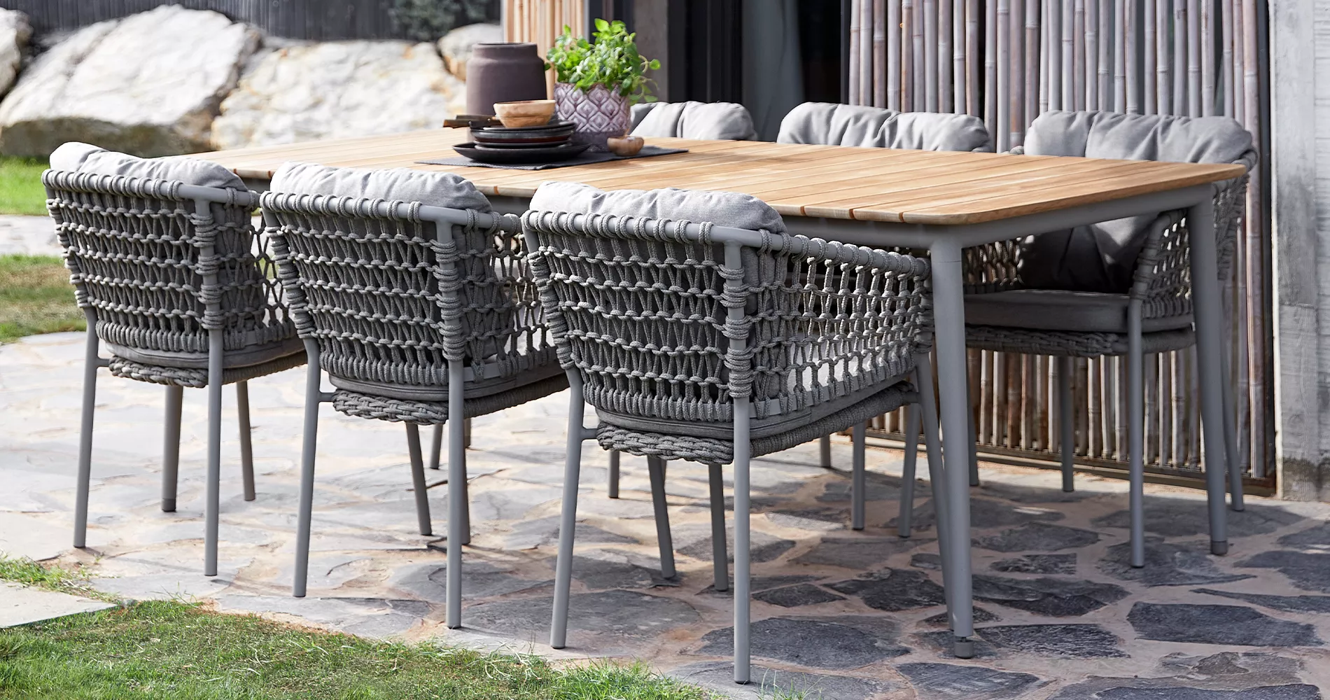 Grau-braune Caneline Ocean Gartenmöbelgruppe auf Steinboden vor Bambuswand, bestehend aus Tisch und acht Stühlen, in natürlicher Umgebung.