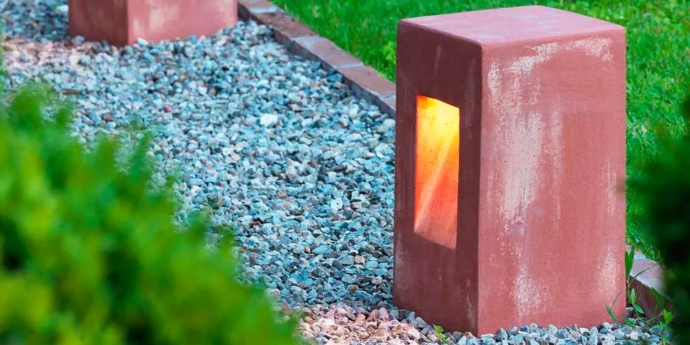 Ethimo Step Outdoor-Leuchte in Rostoptik auf einem Kiesweg neben einer Rasenfläche bei Dämmerung.