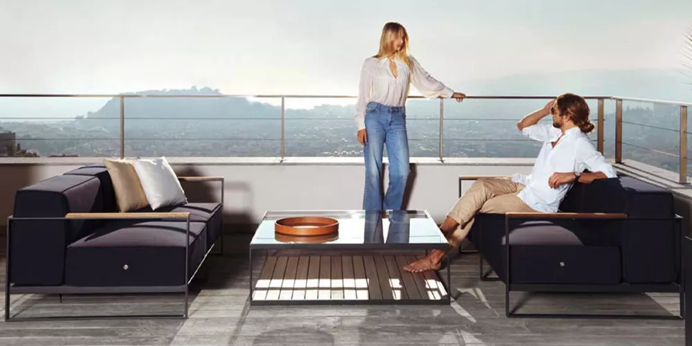 Zwei Personen entspannen auf der Terrasse mit Roshults Monaco Kollektion in Dunkelblau, bestehend aus Sofas und einem Glastisch, vor Bergkulisse.