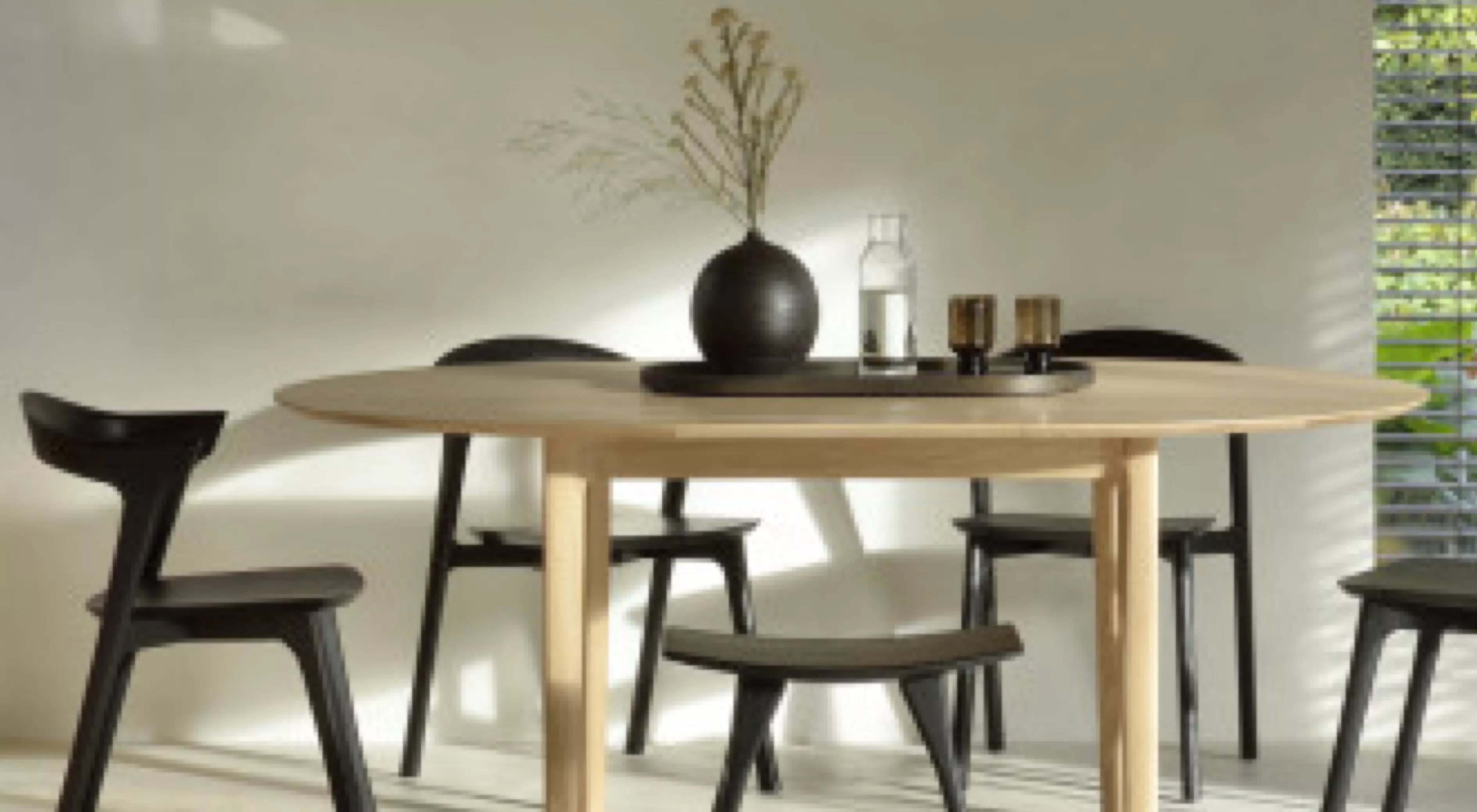 Runder Esstisch aus hellem Holz mit schwarzen Stühlen, dekoriert mit Vase und Gläsern, in einem sonnendurchfluteten Raum.
