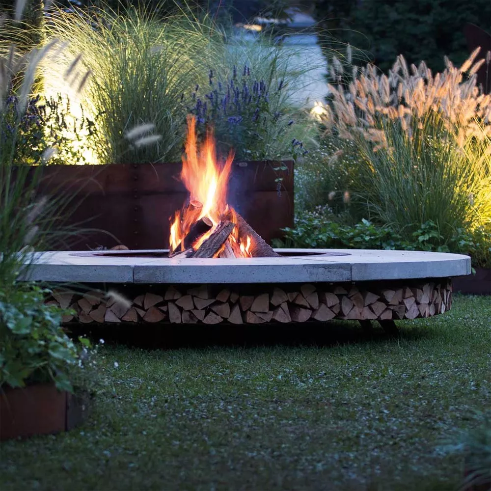 AK47 Ercole Beton Stahl Feuerstelle im Garten mit brennendem Feuer umgeben von Pflanzen und Gräsern in einer stimmungsvollen Abendatmosphäre