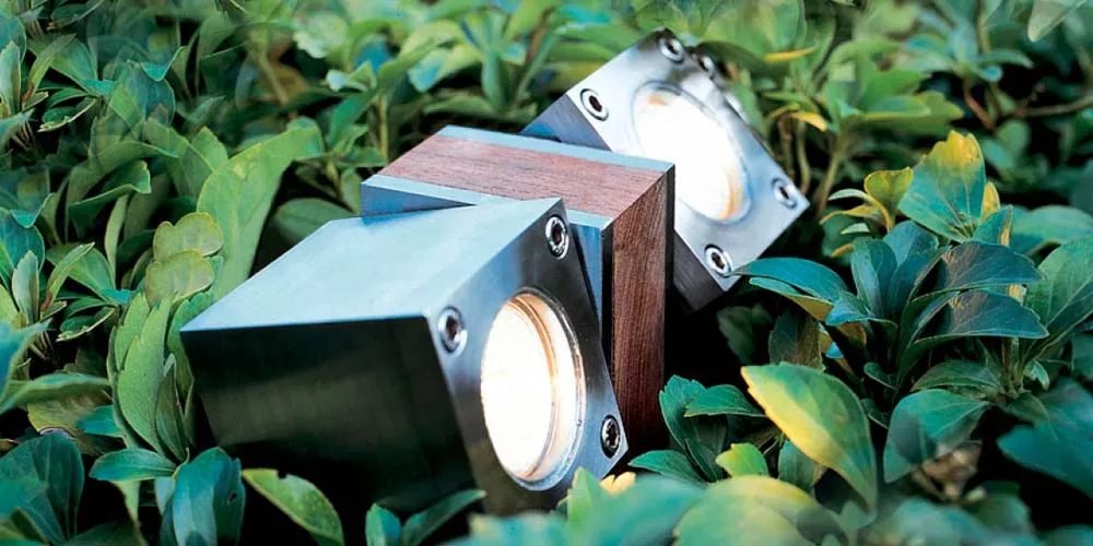 Royal Botania Q-Bic Gartenleuchten in Edelstahl und Holz, eingebettet in grünes Laub, stimmungsvolles Gartenbild.