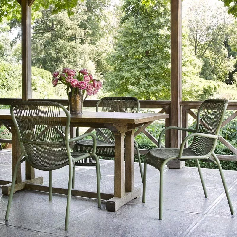 Zwei grüne Vincent's Garden Loop Gartenstühle auf einer Terrasse mit Holztisch und Blumenstrauß, umgeben von üppigem Garten.