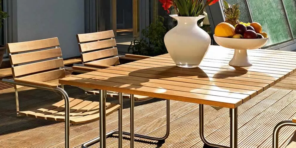 Weißhäupl Prato Gartenmöbel-Set aus Holz auf einer sonnigen Terrasse mit Obstschale und Blumenvase im Hintergrund.