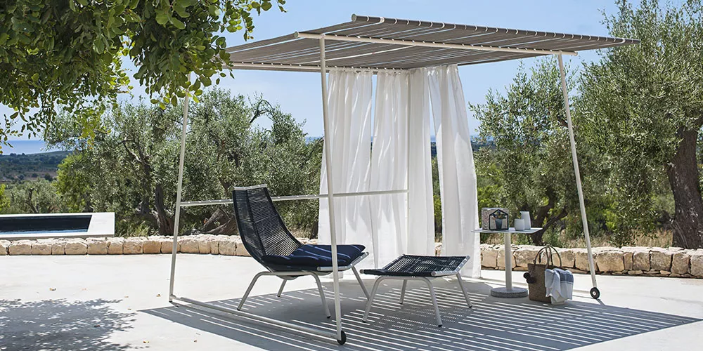 Roda Kollektion Ombria, weißes Sonnendach mit Vorhängen, schwarzer Liegestuhl und Tisch auf Terrasse mit Olivenbäumen im Hintergrund.